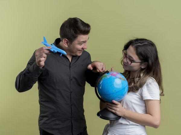 happy young couple of tourists man and woman holding globe and toy air plane standing together having fun over light background